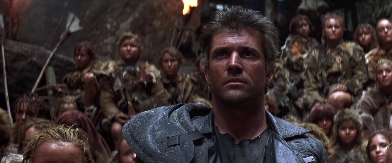 Mad Max Beyond Thunderdome (1985) movie still: medium — A man stands in a dark, rocky enclosure surrounded by a crowd of children. The group wears…