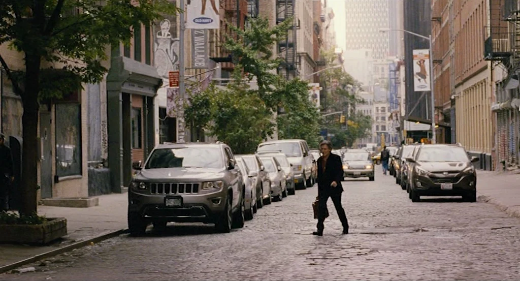 Louder Than Bombs (2015) movie still: wide — A city street paved with weathered cobblestones is lined with parked cars and buildings. An elderly man…