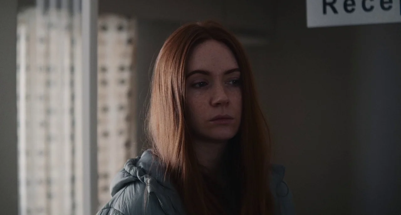 Dual (2022) movie still: closeup — A reception area with a sign visible on the wall and a patterned curtain. A woman with long red hair stands in…