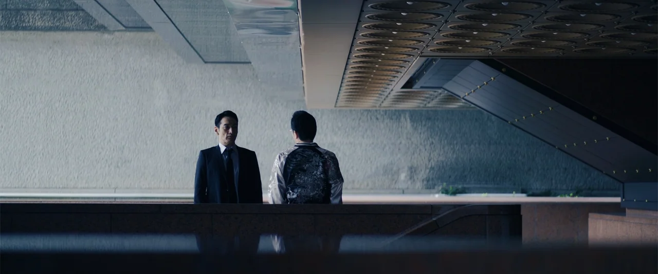 Angel Flight THE MOVIE (2026) movie still: medium — A modernist concrete structure features a low wall and a heavy, repeating ceiling pattern. Two…