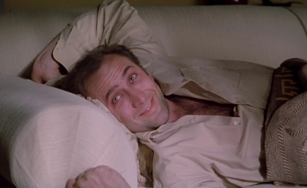 Leaving Las Vegas (1995) movie still: closeup — A man rests on a beige upholstered sofa, leaning against a matching rounded pillow. The man is…