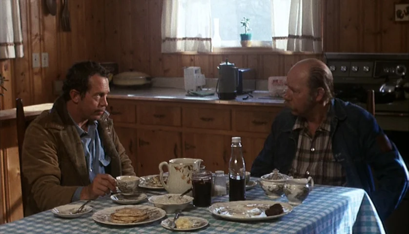 Cockfighter (1974) movie still: medium — A rustic wood-paneled room features a table set for breakfast with various plates. Two men sit opposite…