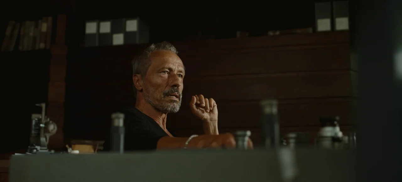 Nope (2022) movie still: medium — A man sits at a desk in a dark room lined with shelves. He glances toward the right while resting his arm on the…