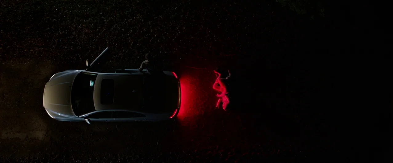 Ghost Stories (2017) movie still: wide — A dark, unpaved rural road at night is lit by the glow of red taillights. A sedan is parked with an open…