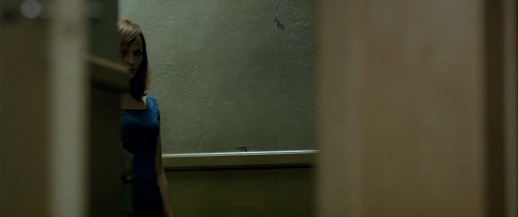 Next Door (2005) movie still: medium — A dimly lit corridor with plain green walls and a horizontal trim strip. A woman stands in the shadows…