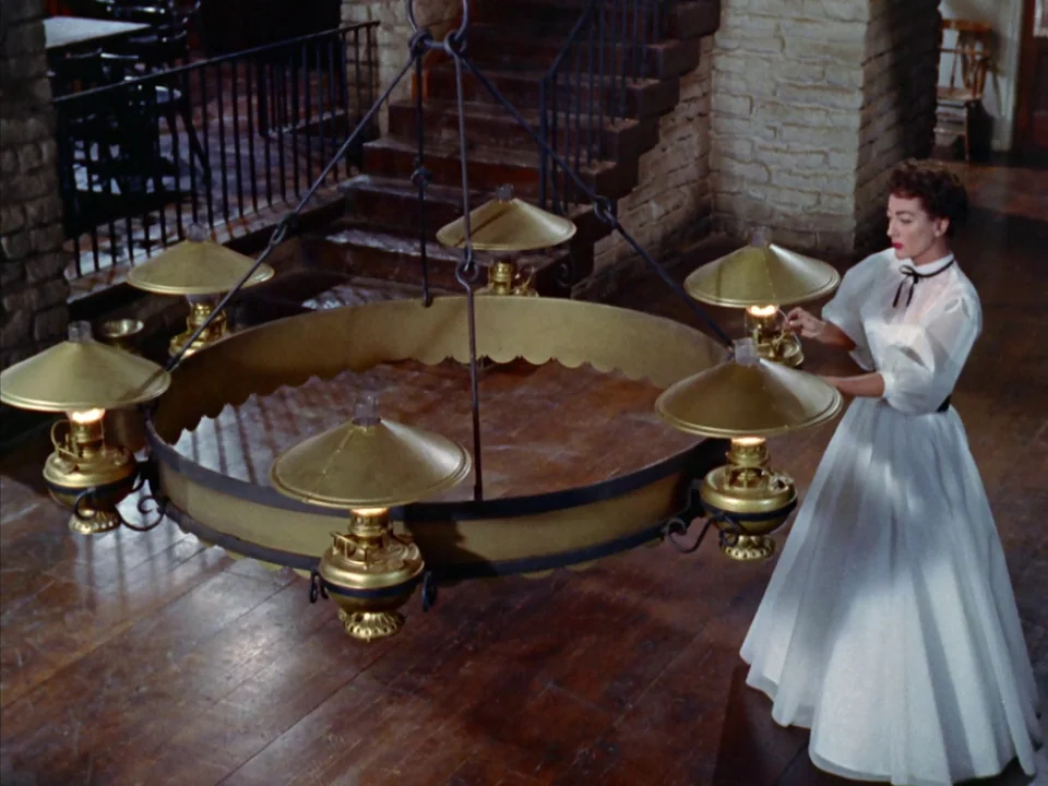 Johnny Guitar (1954) movie still: fullbody — A rustic hall with brick walls and wooden stairs features a large, low-hanging chandelier. A woman in a…