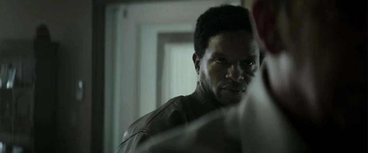 Crown Heights (2017) movie still: medium — A dimly lit residential interior features a person partially obscured in the foreground right. A man in a…