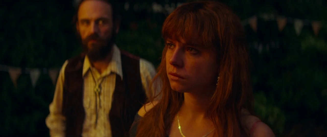 Wild Rose (2018) movie still: medium — Two individuals stand closely together outdoors at dusk with dark foliage and bunting behind. The woman looks…