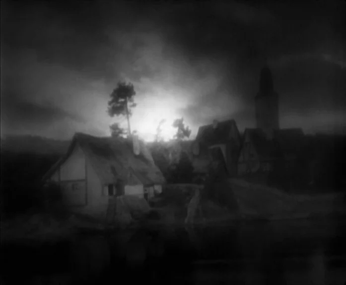 Sunrise (1927) movie still: establishing — A small medieval village sits on the bank of a river under a dark sky. The silhouette of thatched-roof…
