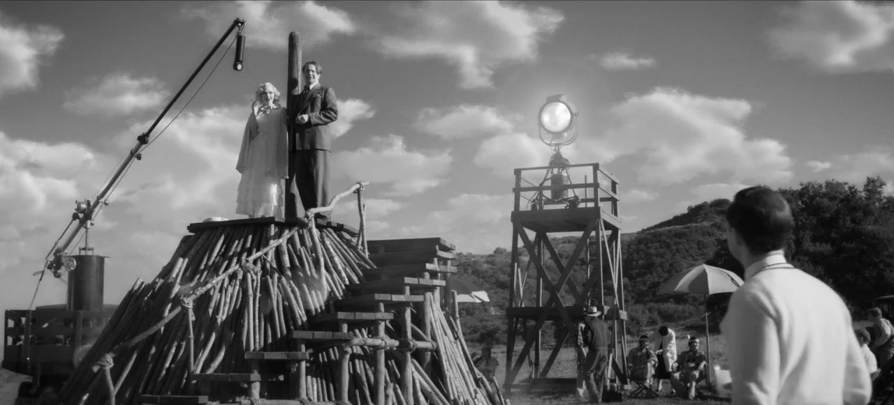 Mank (2020) movie still: wide — A film production set features a large wooden platform constructed from logs and beams. A man and a woman stand atop…