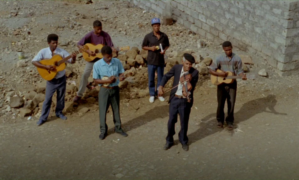 Casa De Lava (1994) movie still: wide — An open dirt lot with piles of rocks and a concrete block wall background. Six men stand in a group playing…