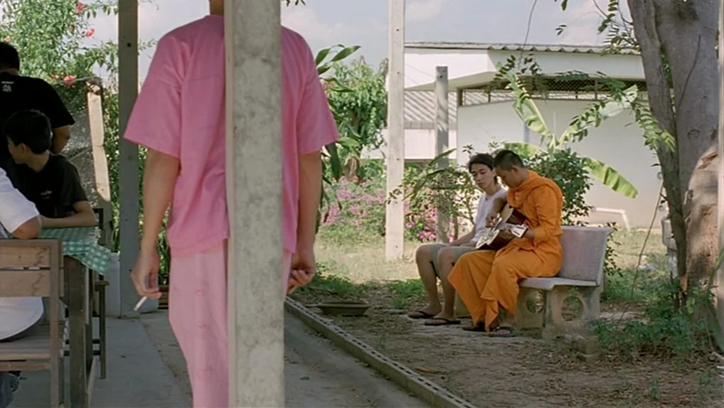 Syndromes and a Century (2006) movie still: medium — An outdoor covered space with concrete pillars features a paved area beside a dirt plot. A…
