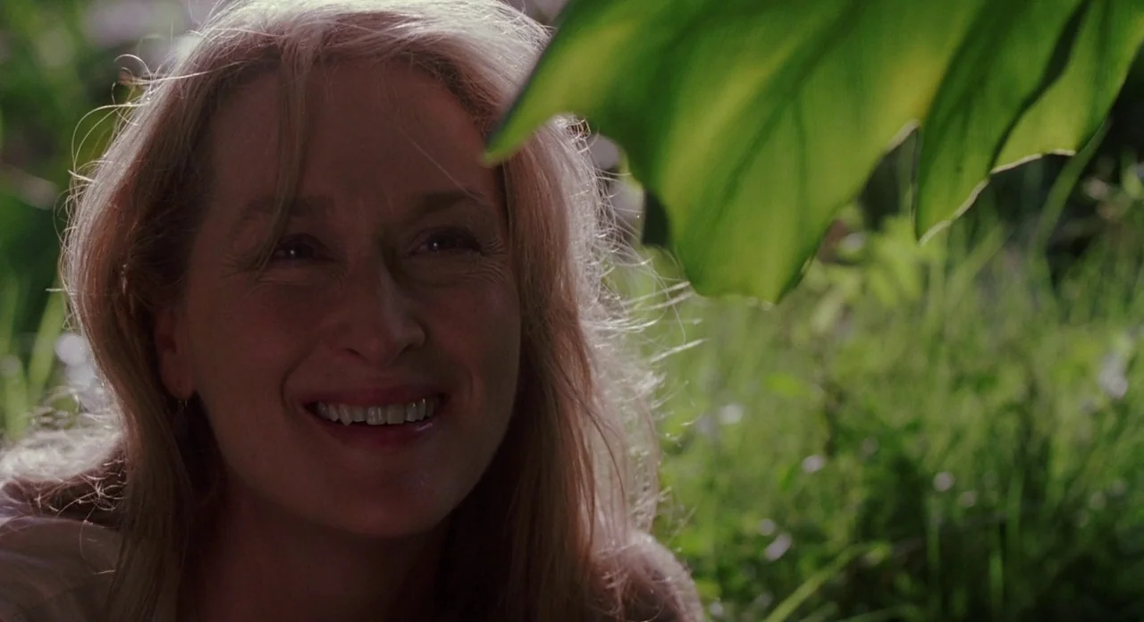 Adaptation (2002) movie still: closeup — A woman stands in a green, leafy garden with sunlight filtering through the trees. She looks towards the…