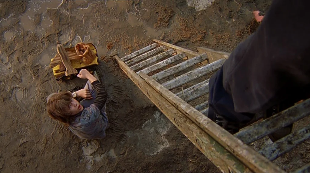 Undertow (2004) movie still: wide — A muddy construction or excavation site with soft earth and puddles. A young boy stands on the ground while an…