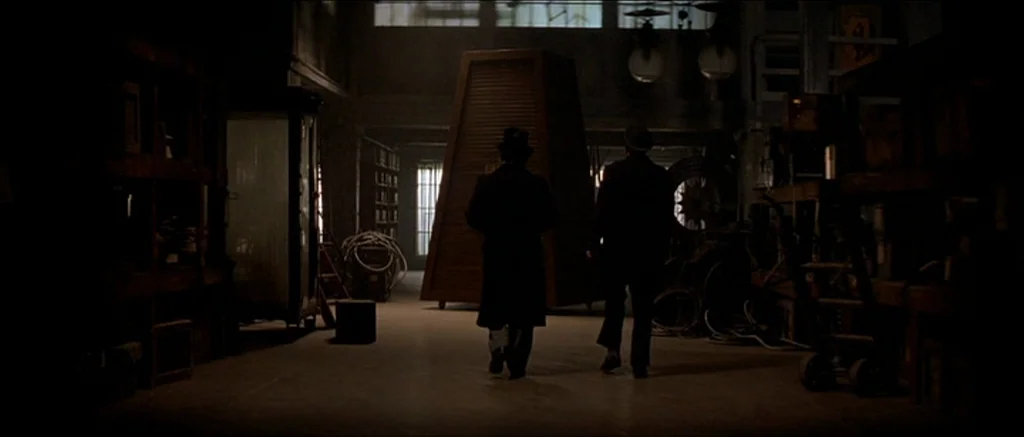 The Prestige (2006) movie still: wide — A dimly lit, cluttered industrial space contains wooden crates and various pieces of equipment. Two men in…