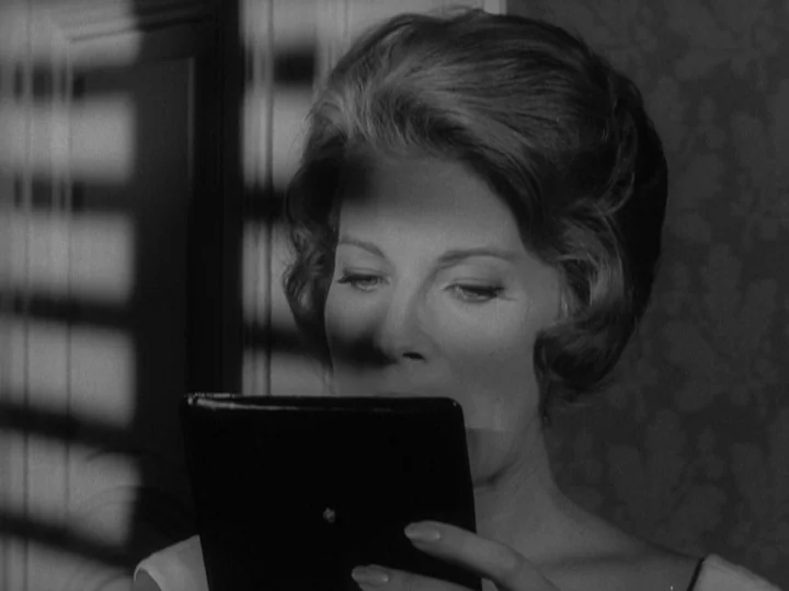 The Silence (1963) movie still: closeup — A woman stands in a room with patterned wallpaper near a slatted window. She holds a small, rectangular…