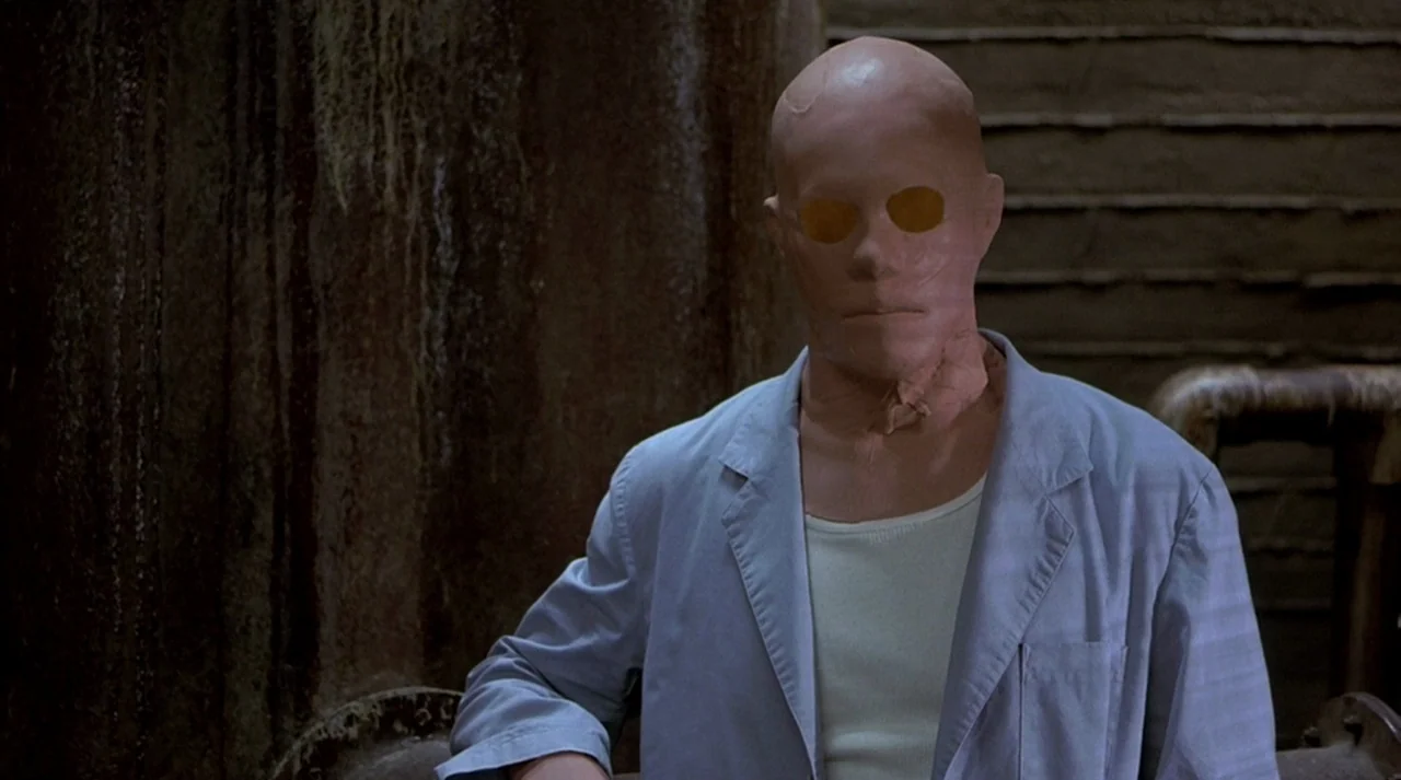Hollow Man (2000) movie still: medium — A man with a featureless head sits in front of a large, corroded industrial pipe. He wears a light blue…