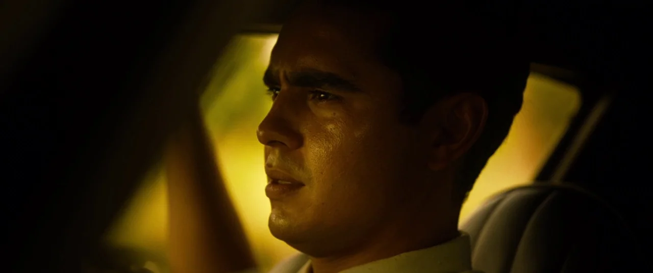 Spiral (2021) movie still: closeup — The interior of a vehicle at night with warm light glowing through the glass. A young man sits in the driver…