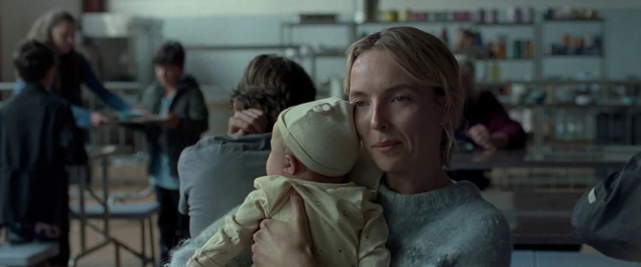 The End We Start From (2023) movie still: medium — A utilitarian institutional space contains rows of tables and metal shelving units in background.…