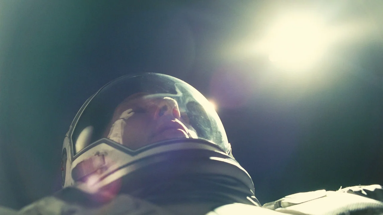 Interstellar (2014) movie still: closeup — A figure in a space suit lies against a dark background. The bright sun casts a glare across the helmet…