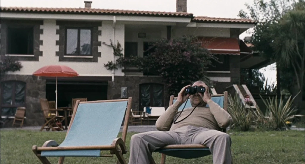 Timecrimes (2007) movie still: medium — A suburban house with a tiled roof stands behind a grassy lawn area. An elderly man sits on a blue lawn…