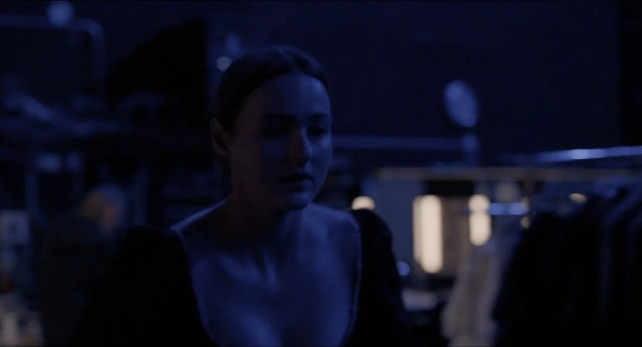 Sentimental Value (2025) movie still: medium — A dimly lit room contains racks of clothing visible in the blurred background. A woman stands in the…