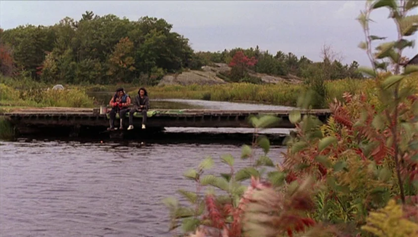 Dance Me Outside (1994) movie still: wide — A wooden dock extends into a calm river bordered by dense green and autumn trees. Two individuals sit…