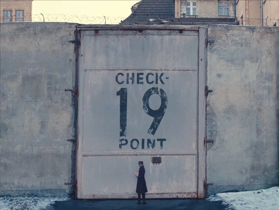 The Grand Budapest Hotel (2014) movie still: wide — A tall concrete wall features a large, industrial sliding gate marked with bold black lettering.…