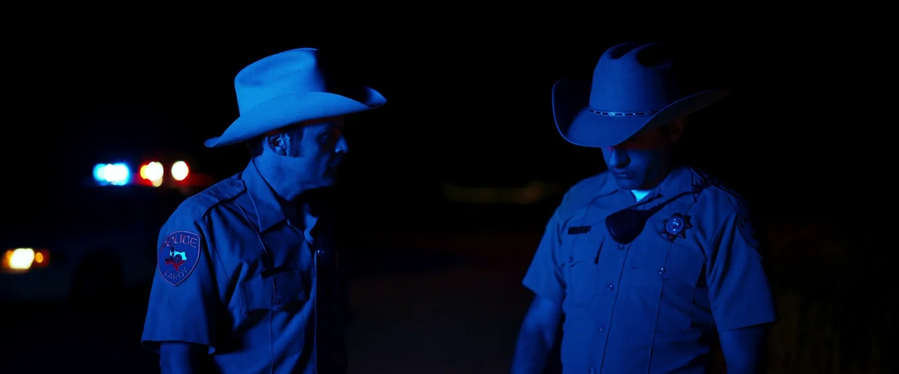 LaRoy, Texas (2023) movie still: medium — Two police officers stand in the darkness illuminated by flickering red and blue lights. A blurred…