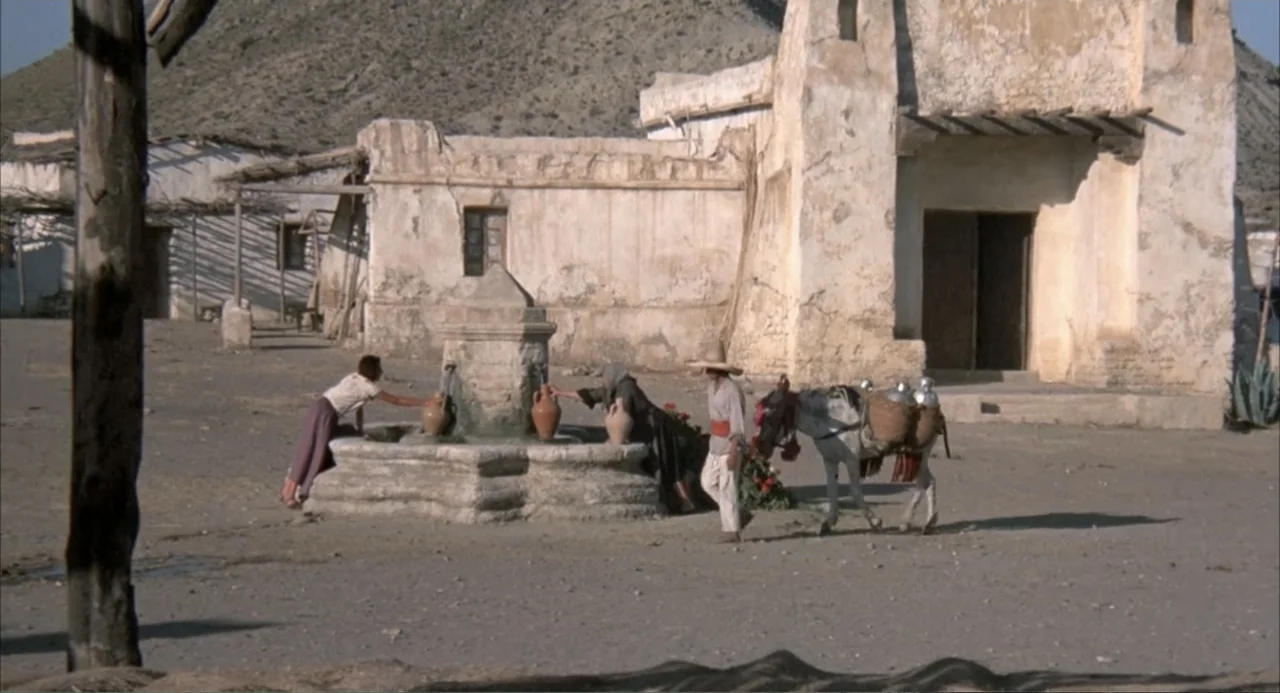 The Spikes Gang (1974) movie still: wide — A sun-baked village square features a central stone fountain and a weathered stucco building. Two women…