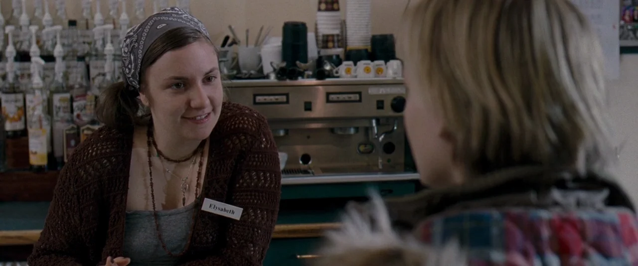 The Innkeepers (2011) movie still: medium — A coffee shop interior features a stainless steel espresso machine and rows of syrup bottles. A woman…