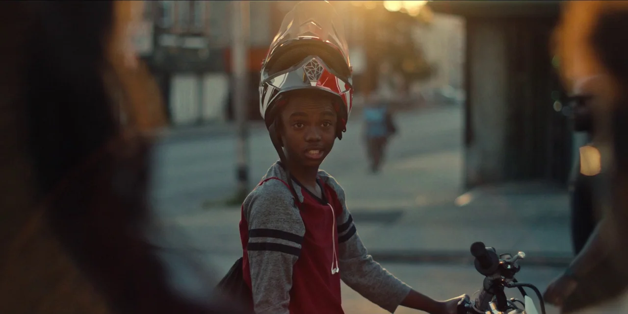 Charm City Kings (2020) movie still: medium — A city street at golden hour with blurred background buildings and a paved road. A young teenage boy…