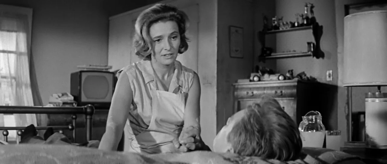 Hud (1963) movie still: medium — A 1960s bedroom features a bedside television, a desk lamp, and a wooden chest. A woman sits on the bed holding the…