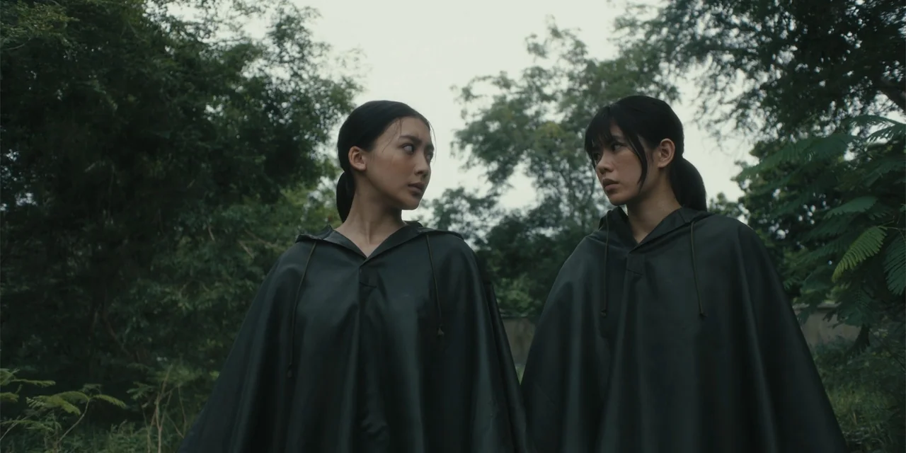 Host (2020) movie still: medium — A dense grove of trees with green foliage surrounds two people in dark ponchos. The two women stand together and…