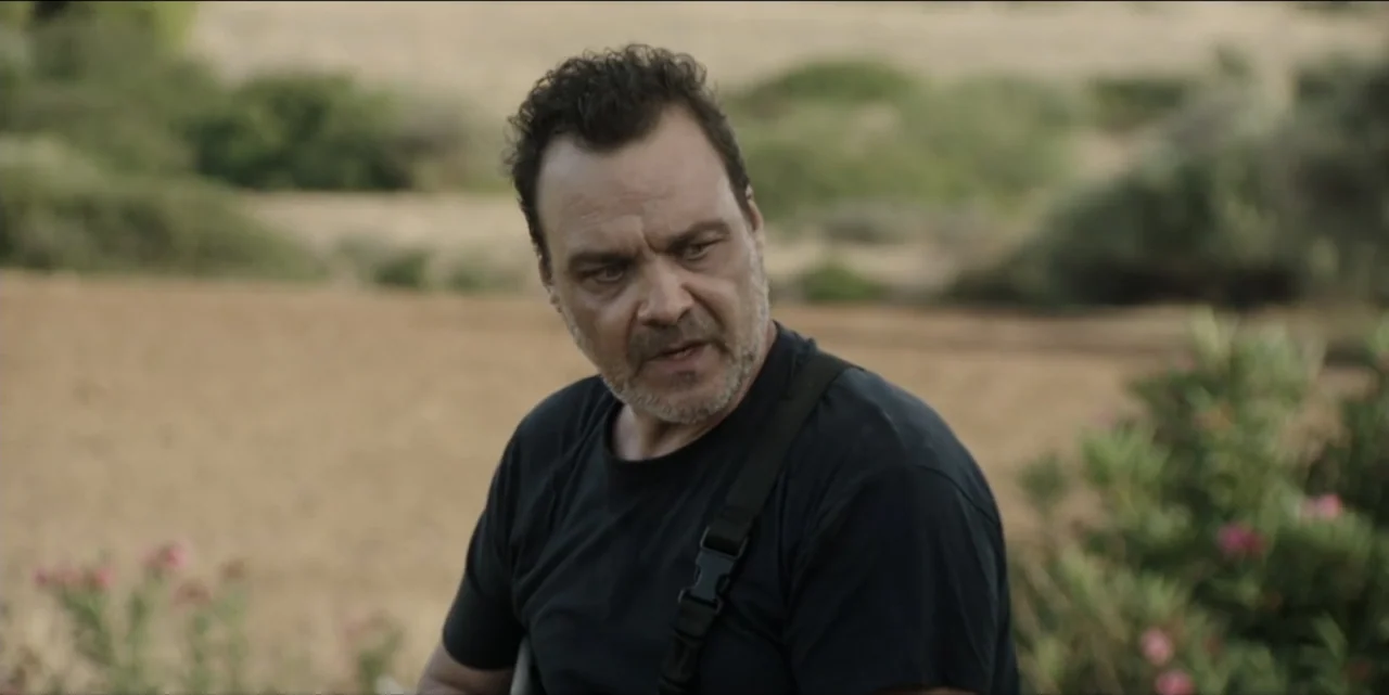 The Hunted (2024) movie still: medium — A man stands outdoors in a dry, open field during the daytime. He is wearing a black t-shirt and looking…