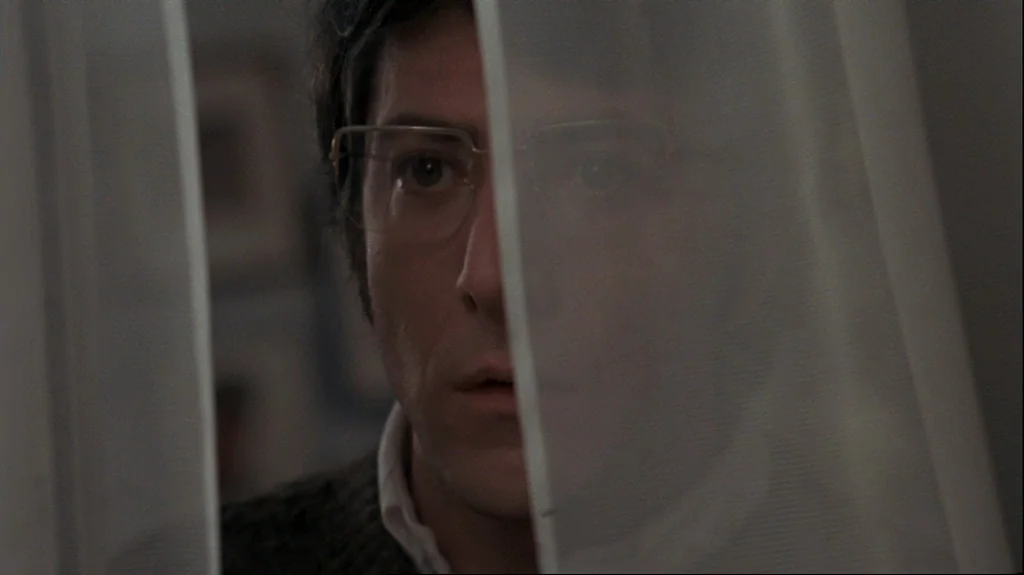 Straw Dogs (1971) movie still: closeup — A man's face appears through the vertical gap of a semi-transparent white curtain. The foreground fabric…