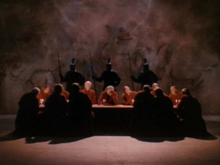 The Magic Flute (1975) movie still: wide — A large room with a textured mural on the back wall depicting primitive hunts. A group of robed figures…