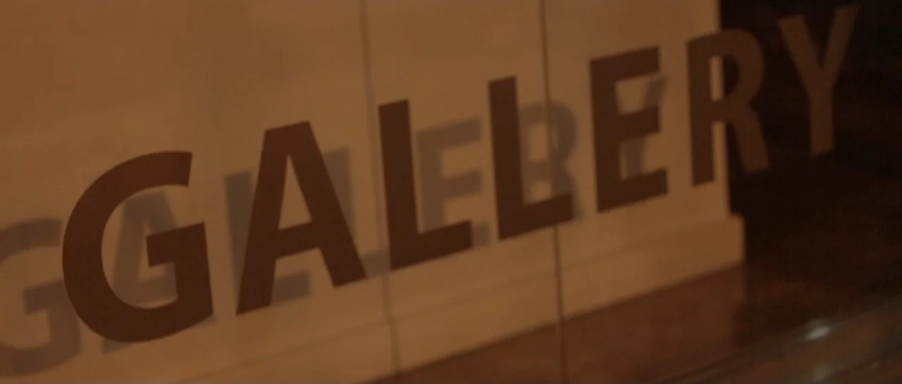 NY84 (2016) movie still: closeup — A cream-colored wall surface displays the word gallery in bold dark brown lettering. The camera captures a close…