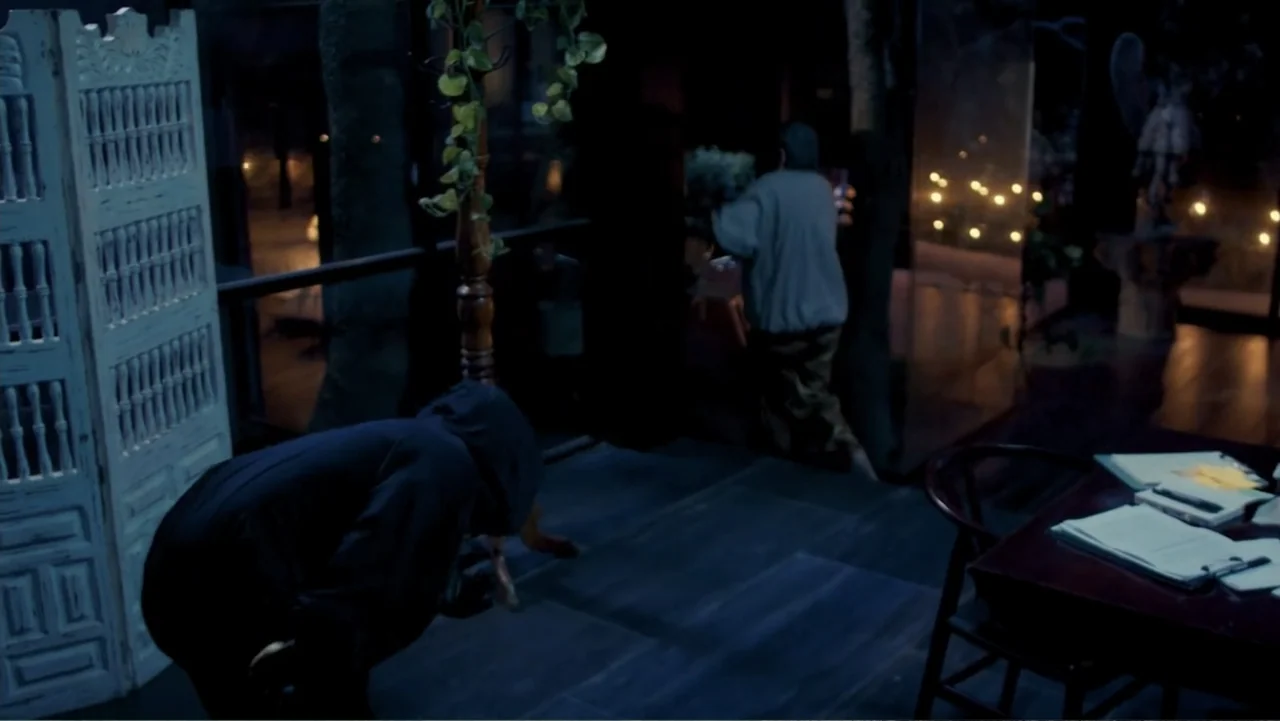 Por Amor al Money (2023) movie still: medium — A dark interior space features a white room divider and a wooden writing desk. One person in a dark…