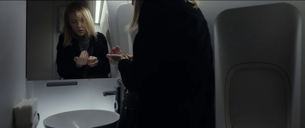 Tár (2022) movie still: medium — A compact airplane lavatory features white panels and a modern chrome faucet. A blonde woman in a black jacket…