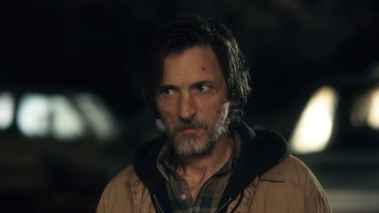 Winter’s Bone (2010) movie still: closeup — A man stands outdoors at night with blurred lights in the background. The man looks toward the side of…