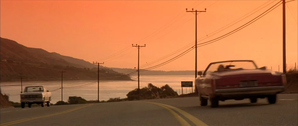 True Romance (1993) movie still: wide — A coastal highway at sunset with a body of water and distant hills visible. A pink convertible and a…