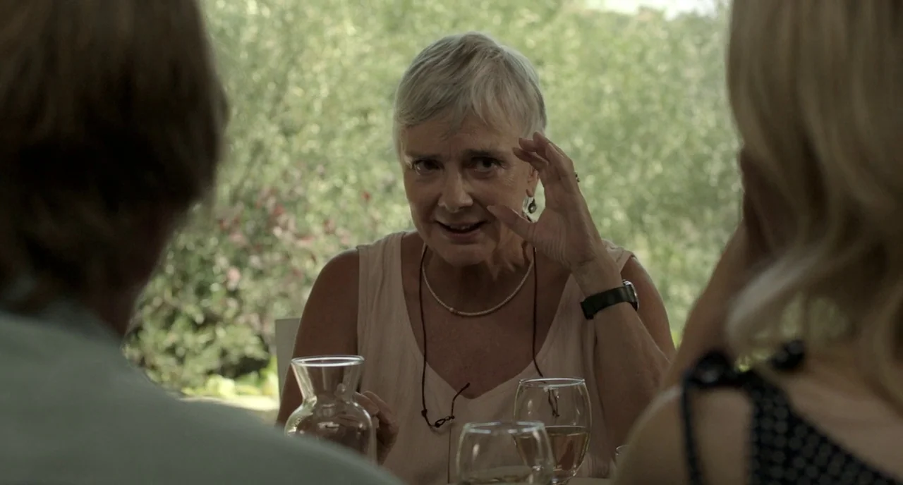 Before Midnight (2013) movie still: medium — An outdoor patio area features lush greenery blurred in the background. An elderly woman gestures with…