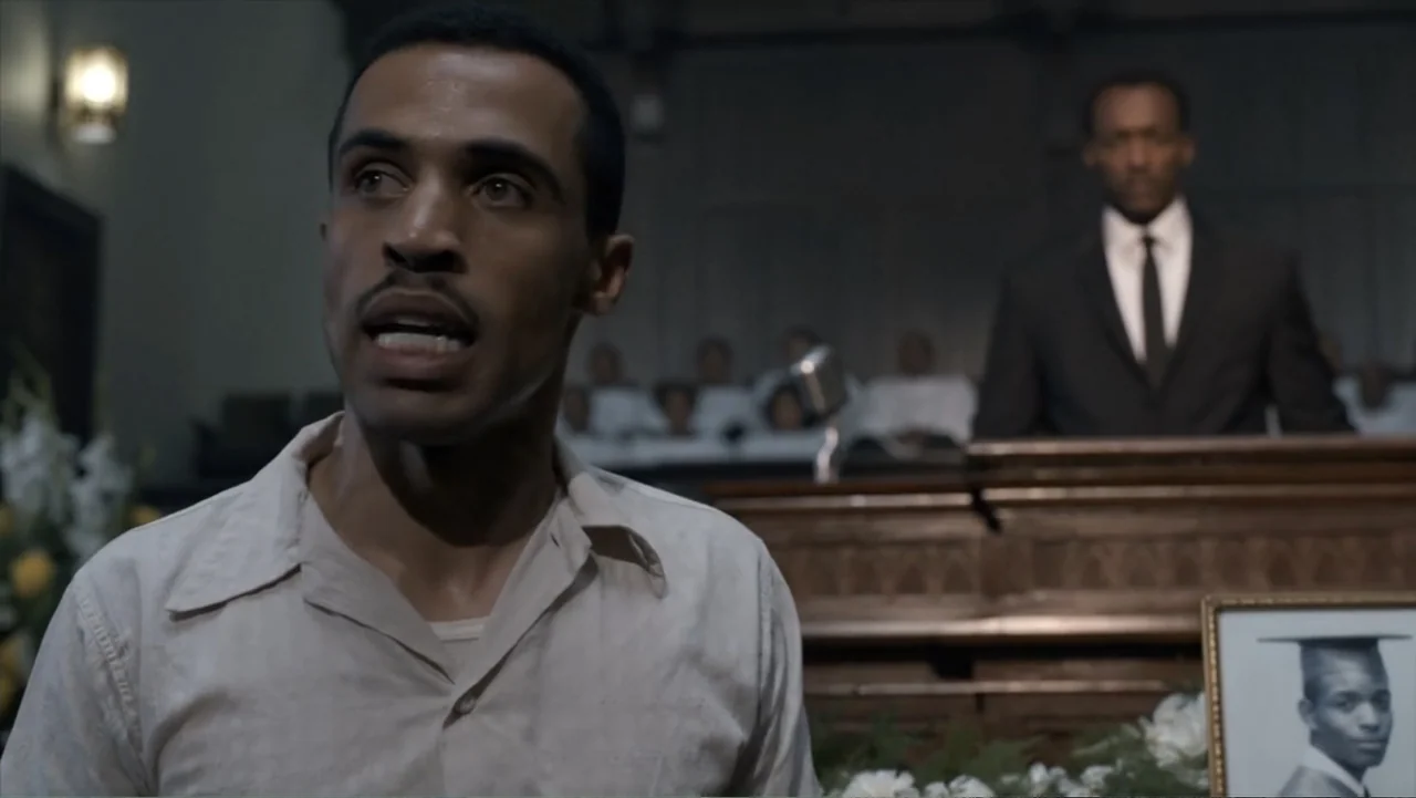All the Way (2016) movie still: medium — A dark church interior contains a wooden pulpit and white floral funeral arrangements. A man speaks in the…