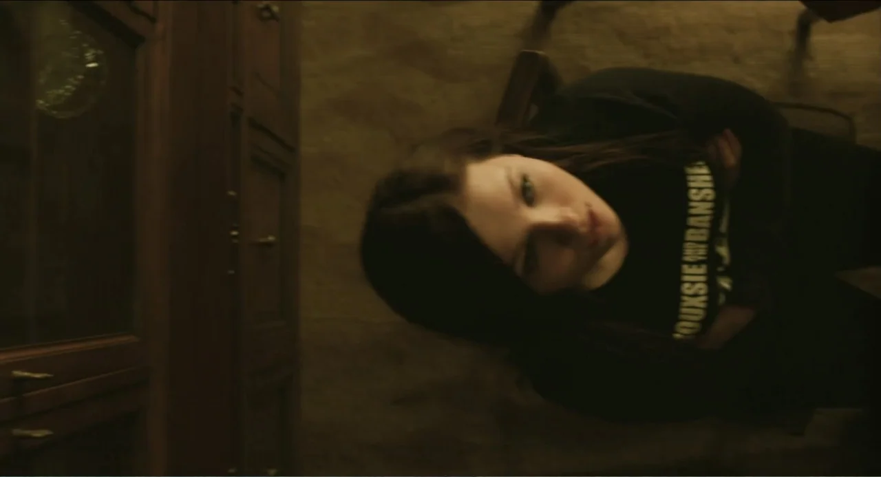 Haunter (2013) movie still: medium — A dimly lit room with dark wooden furniture and a carpeted floor. A young woman in a black t-shirt lies on a…