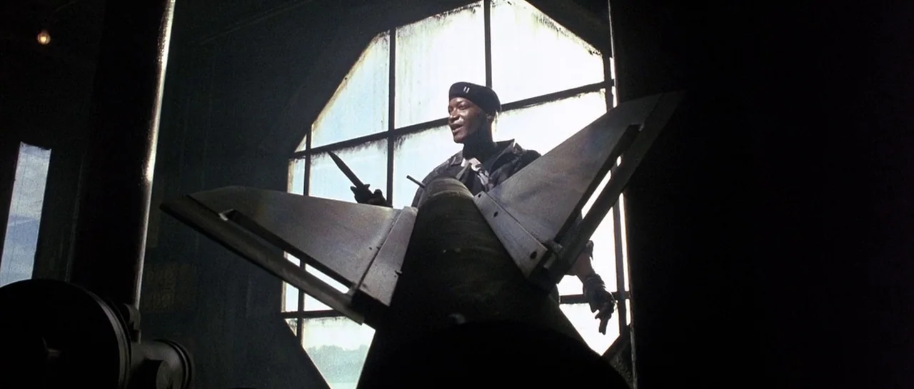 The Rock (1996) movie still: medium — A dimly lit industrial space features a large octagonal window behind a missile. A man in a beret stands…
