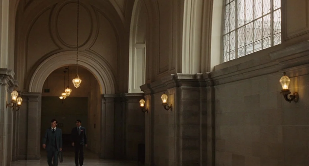 Milk (2008) movie still: wide — A stone corridor with high vaulted ceilings and large windows on one side. Two men walk down the hallway together…