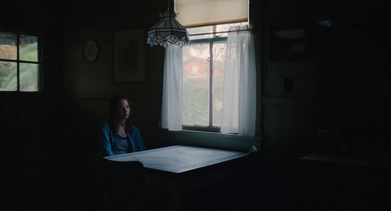 Causeway movie still: medium — A dimly lit room features a worn table positioned near a curtained window. A woman sits quietly at the table while…