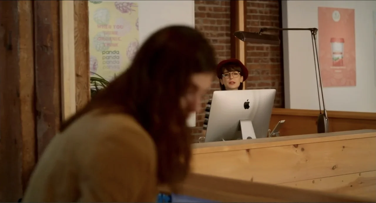 Can You Keep a Secret? (2019) movie still: medium — An interior space features a wooden front desk and a brick wall background. A woman sits behind…