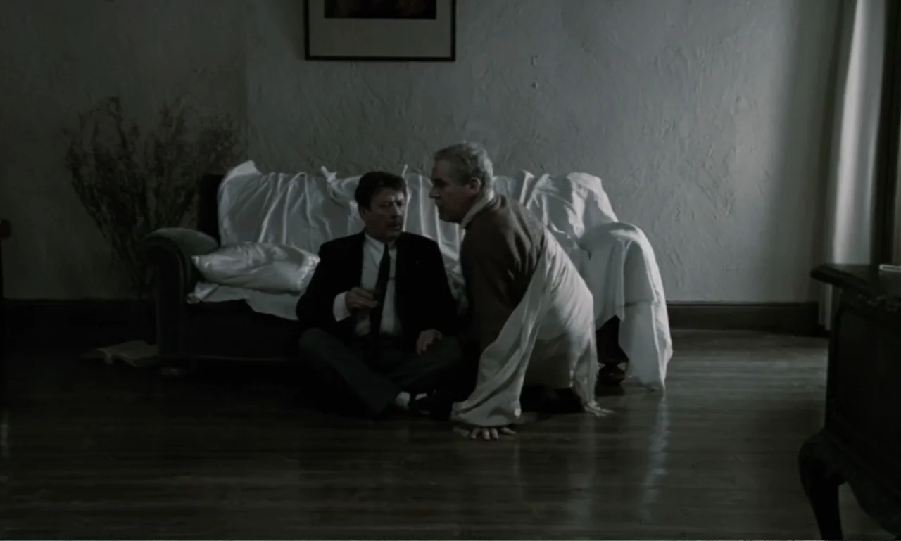 The Sacrifice (2015) movie still: medium — A sparsely furnished living room contains a couch covered with a white sheet. One man sits on the wooden…